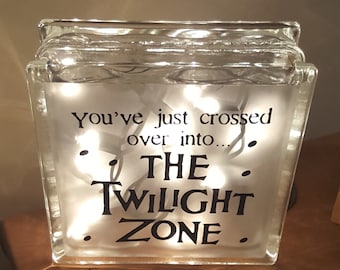 May include: A glass block with a white background and the text "You've just crossed over into... THE TWILIGHT ZONE."  The block is illuminated with white fairy lights.