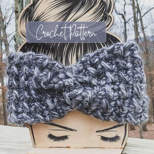 Könnte beinhalten: Ein grobes, gedrehtes Häkelstirnband aus einem melierten Garn in Anthrazit, Schwarz und Weiß. Das Stirnband wird auf einem hölzernen Kopfmodell präsentiert, das mit dem Text Crochet Pattern in weißer Schreibschrift versehen ist.