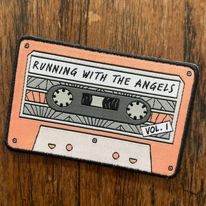 May include: A pink and white cassette tape patch with black stitching. The tape has a black and gray cassette tape design with the text "RUNNING WITH THE ANGELS VOL. 1" printed on it.