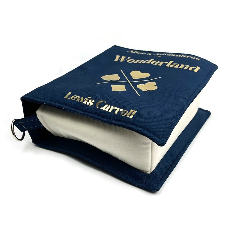 May include: A blue canvas book-shaped storage box with a white interior. The box has gold lettering that reads "Alice's Adventures in Wonderland" and "Lewis Carroll".