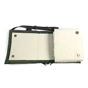 May include: A green canvas crossbody bag with a brown strap and a flap that opens to reveal a white canvas interior. The bag has a zipper closure and a small pocket on the front.