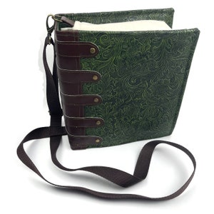 May include: A green faux leather book-style bag with a brown leather strap and metal accents. The bag has a floral embossed design and a long adjustable shoulder strap.
