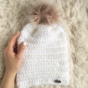 Puede incluir: Gorro blanco de punto de ganchillo con un pomp&oacute;n marr&oacute;n esponjoso y una etiqueta de cuero marr&oacute;n que dice "HORCS".