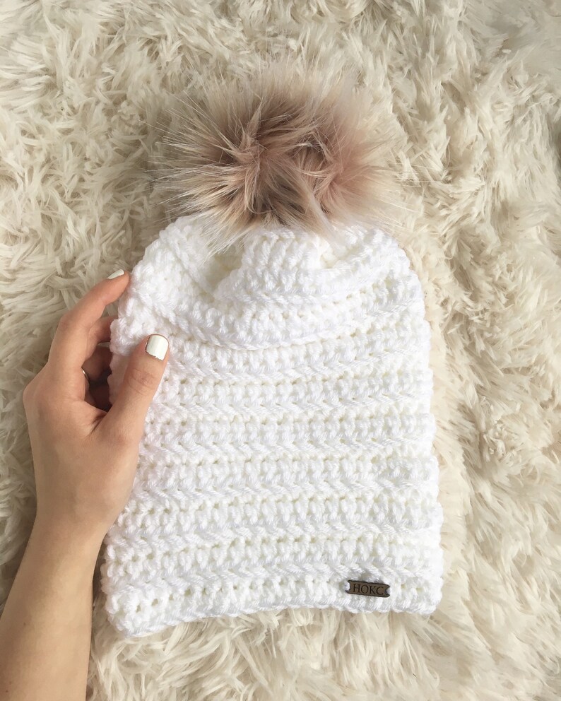 Puede incluir: Gorro blanco de punto de ganchillo con un pomp&oacute;n marr&oacute;n esponjoso y una etiqueta de cuero marr&oacute;n que dice "HORCS".