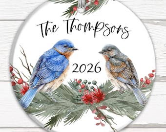 May include: A white round ceramic bauble featuring two bluebirds perched on a branch with green leaves and red berries. The bauble is inscribed with "The Thompsons 2026" in black text.