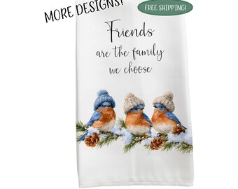 May include: A white kitchen towel featuring the text Friends are the family we choose in black script. Below the text are three bluebirds with orange chests, each wearing a small knit hat, perched on a snowy pine branch with brown pinecones.