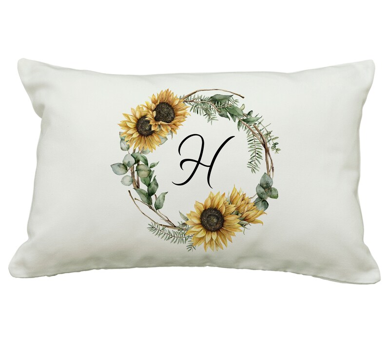 May include: A rectangular white pillow featuring a watercolor design. The design includes a wreath of sunflowers, greenery, and the letter "H" in black script. The sunflowers are yellow and brown.