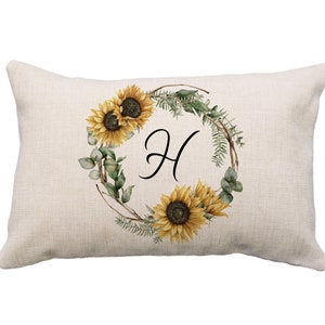 May include: Rectangular beige pillow with a watercolor sunflower wreath design. The wreath surrounds the black letter "H". The wreath includes yellow sunflowers, green leaves, and brown branches. The pillow has a textured appearance.