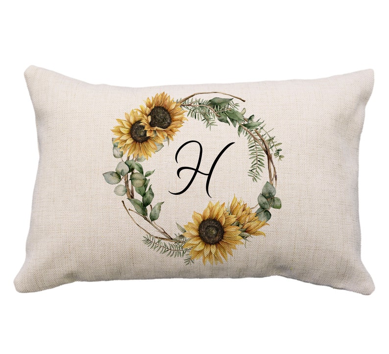May include: Rectangular beige pillow with a watercolor sunflower wreath design. The wreath surrounds the black letter "H". The wreath includes yellow sunflowers, green leaves, and brown branches. The pillow has a textured appearance.