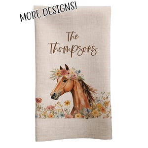 May include: A beige linen-textured towel featuring a watercolor illustration of a horse wearing a floral crown. The text "The Thompsons" is above the horse image. The words "MORE DESIGNS!" are in the upper left corner.