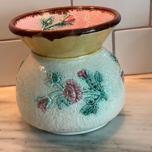 May include: A ceramic vase with a wide, flared rim featuring a brown edge and a pink interior. The exterior has a textured, off-white finish decorated with raised, molded floral motifs in shades of pink and green, encircling the rounded body.