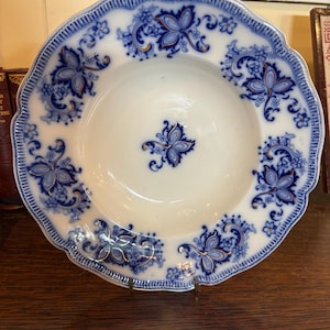 May include: A white ceramic plate with a scalloped edge and a blue floral pattern. The plate features a central floral design and repeating floral motifs around the rim. Gold accents highlight the blue and white design. The plate is displayed on a metal stand.