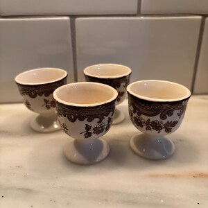 May include: A set of four cream-colored ceramic egg cups featuring a dark brown transferware pattern. Each cup has a pedestal base and a decorative border of floral motifs and intricate scrollwork around the upper rim of the exterior.