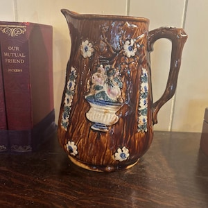 May include: A brown ceramic pitcher with a textured, bark-like appearance. The pitcher features a floral relief design with white, blue, and pink accents. The handle is also textured, and the pitcher is sitting on a wooden surface.