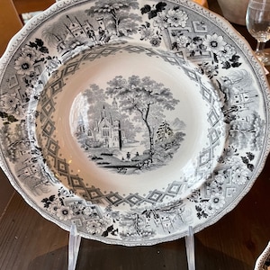 May include: A black and white ceramic soup bowl with a detailed floral and architectural design. The bowl features a central image of a building with trees and figures.