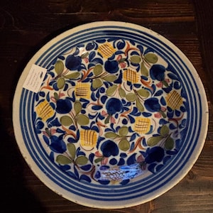 May include: A round, hand-painted ceramic plate with a blue and white striped border. The plate's interior features a floral design with blue, yellow, green, and red accents. The plate is set on a dark wooden surface.