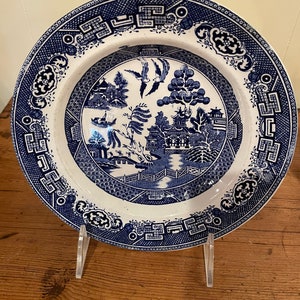 May include: A blue and white porcelain plate with a traditional willow pattern design. The plate features a central image of a pagoda, a willow tree, and two birds in flight. The rim of the plate is decorated with a repeating pattern of blue and white.