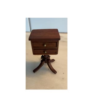 May include: Miniature wooden nightstand with two drawers and brass knobs. The nightstand is dark brown and sits on a decorative base. A small piece of furniture for a dollhouse or other miniature setting.