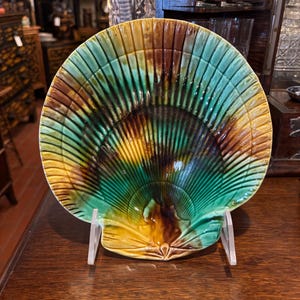 May include: A ceramic dish shaped like a large scallop shell, featuring a glossy glaze in shades of emerald green, golden yellow, and deep brown. The surface is textured with radiating ridges that extend from the base to the scalloped edge.