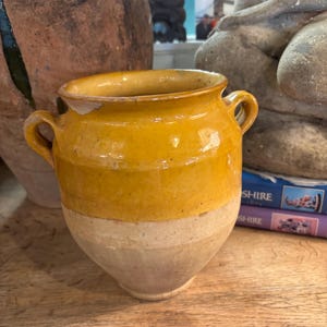 May include: A two-handled earthenware pot featuring a glossy mustard-yellow glazed upper section and a matte, unglazed beige lower body. The vessel has a wide, flared rim with a small chip on the edge and a rounded, tapered base design.