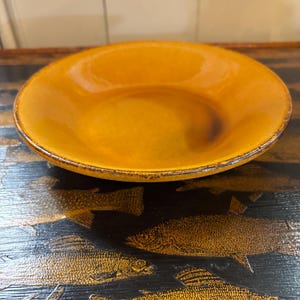 May include: A shallow, circular ceramic bowl with a glossy, warm mustard-yellow glaze. The rim features a dark, distressed brown edge with visible texture. The bowl has a smooth, concave interior surface and a slightly raised, rounded outer edge.
