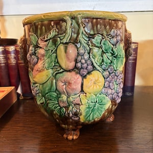May include: A ceramic planter with a detailed relief design of fruits and leaves. The planter features grapes, peaches, and other fruits in shades of green, purple, yellow, and brown. It has a wide opening and three small feet.
