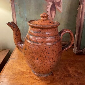 May include: A brown speckled ceramic teapot with a lid. The teapot has a rounded body and a spout on the side. The teapot is sitting on a wooden surface.
