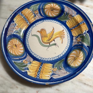 May include: A hand-painted ceramic plate with a blue rim and a central design featuring a yellow bird with a green branch. The plate has yellow and blue floral patterns and measures approximately 10 inches in diameter.