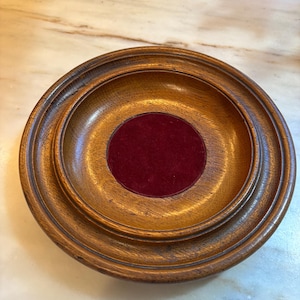 May include: A round, wooden trinket dish with a dark red velvet center. The dish has a tiered design with a raised outer rim and a smooth, polished finish. The wood has a warm, brown tone, and the velvet center provides a contrasting texture.