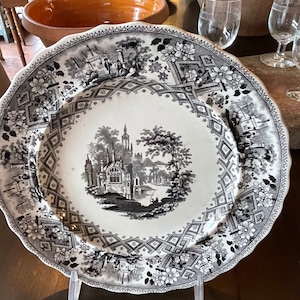 May include: A black and white porcelain plate with a detailed floral border and a central image of a castle and trees. The plate is on a wooden table and is being displayed on a clear plastic stand.