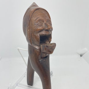 May include: A wooden nutcracker carved in the shape of a person's head wearing a hat. The mouth is open and the jaw is hinged to crack nuts.