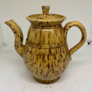 May include: A brown and yellow ceramic teapot with a speckled glaze. The teapot has a round body and a spout on one side and a handle on the other.