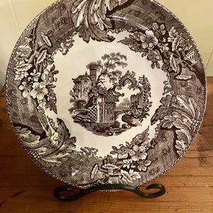 May include: A brown and white ceramic plate with a floral design and a central image of a building with a garden. The plate is on a black stand.