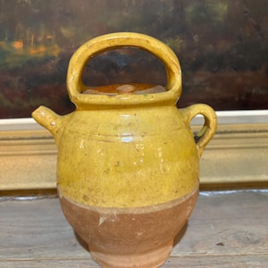May include: A small, yellow ceramic teapot with a brown base and handle. The teapot has a spout and a small lid. The background includes a painting and a wooden surface.