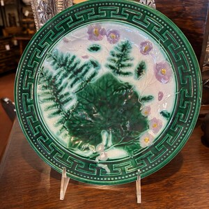 May include: A round ceramic plate featuring a dark green border with a Greek key pattern. The center displays a white background with embossed green fern leaves, a large central green leaf, and small light purple flowers with yellow centers.
