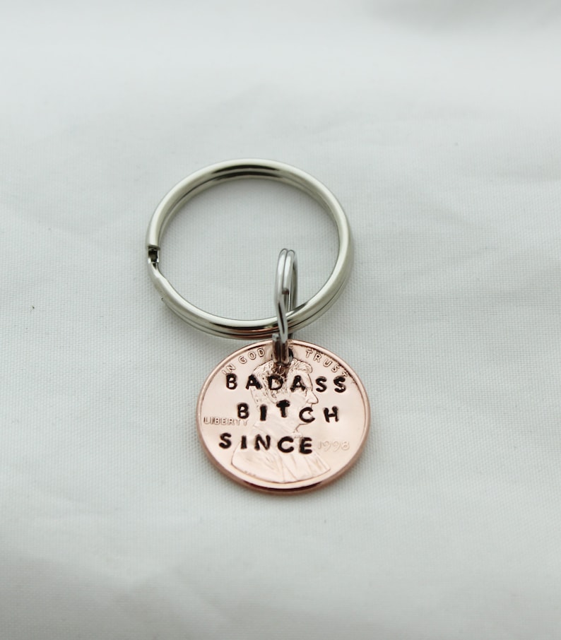 May include: A copper-colored penny keychain with the words "Badass Bitch Since" engraved on it. The keychain is attached to a silver key ring.