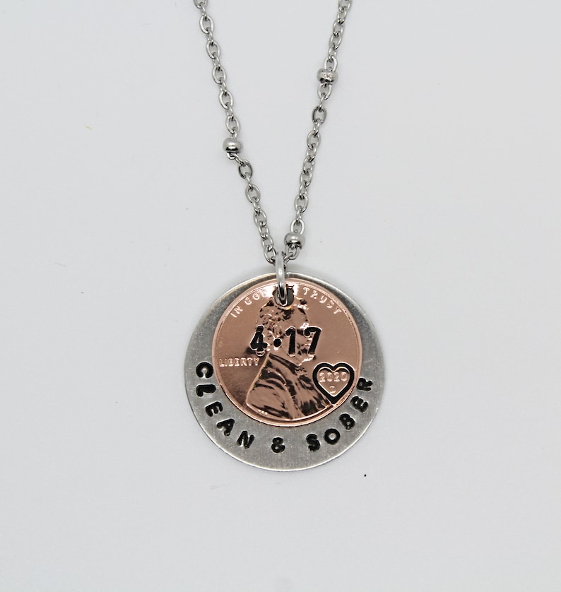 May include: A silver necklace with a copper penny pendant. The penny has the date 4-17 and a heart with the year 2020. The pendant is surrounded by a silver circle with the words "Clean & Sober" engraved on it.