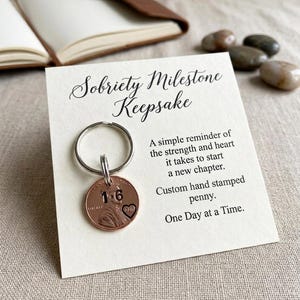 May include: A copper-colored penny keychain attached to a white card with black text that reads Sobriety Milestone Keepsake, A simple reminder of the strength and heart it takes to start a new chapter. Custom hand stamped penny. One Day at a Time.