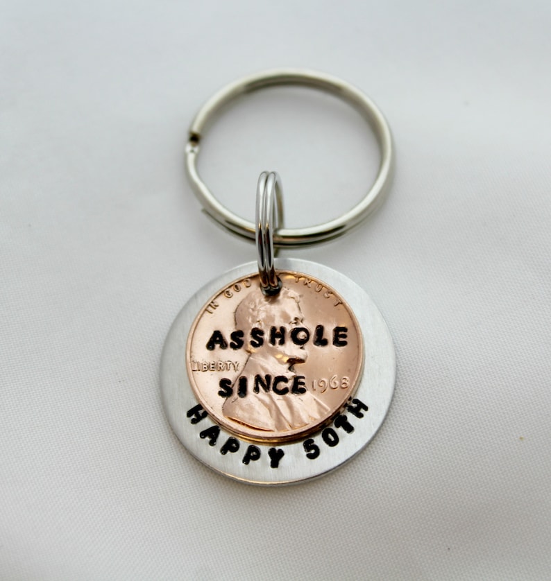 May include: A silver key chain with a copper penny attached. The penny has the text "ASSHOLE SINCE 1968" on it. The keychain has the text "HAPPY 50TH" on it.
