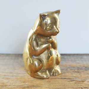 May include: A solid brass figurine of a cat sitting with its paws tucked in. The cat is a light gold color and has a detailed face.