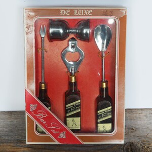 May include: A vintage De Luxe bar set with a bottle opener, two shot glasses, a bottle opener, and a bottle of Johnnie Walker Black Label Scotch Whisky. The set is packaged in a red and white box with the words "Bar Set" printed on it.