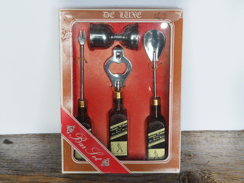 May include: A vintage De Luxe bar set with a bottle opener, two shot glasses, a bottle opener, and a bottle of Johnnie Walker Black Label Scotch Whisky. The set is packaged in a red and white box with the words "Bar Set" printed on it.
