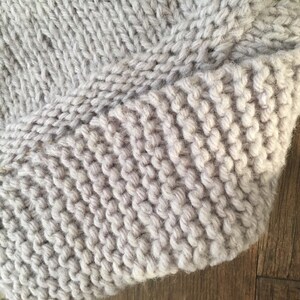 May include: Close-up of a light gray knitted fabric with a textured surface. The fabric is folded over on itself, showing the knit stitches.