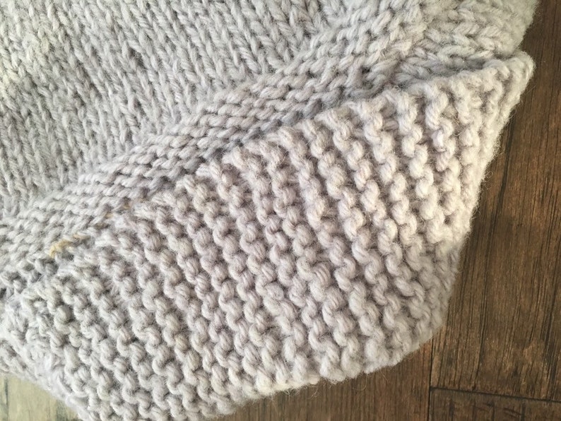 May include: Close-up of a light gray knitted fabric with a textured surface. The fabric is folded over on itself, showing the knit stitches.