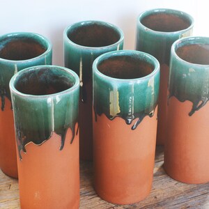 Pode incluir: Seis xícaras de cerâmica de terracota com esmalte verde e detalhes pretos. As xícaras estão dispostas em fila sobre uma superfície de madeira.