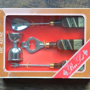 May include: A silver bar set in a brown box with the text "De Luxe Bar Set". The set includes a bottle opener, a jigger, a spoon, and a cocktail stick.
