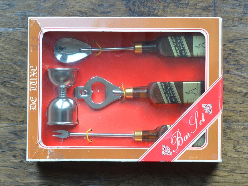 May include: A silver bar set in a brown box with the text "De Luxe Bar Set". The set includes a bottle opener, a jigger, a spoon, and a cocktail stick.