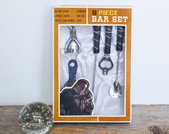 Op de afbeelding: Een vintage 6-delige barset in een bruine en witte doos. De set bevat een roerlepeltje, een zeef, een dubbele maatbeker, een baraccessoire, een fles- / blikopener en een prikker.