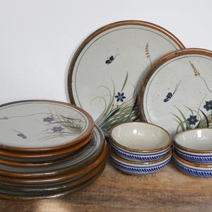 May include: Set of ceramic dinnerware featuring a floral design. The set includes a stack of plates and two bowls with blue and white trim.