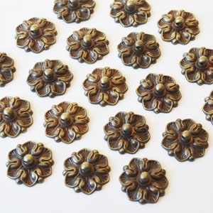 May include: A collection of 18 antique gold-toned metal flower-shaped embellishments with a raised centre. The embellishments have a detailed design and a slightly distressed finish.